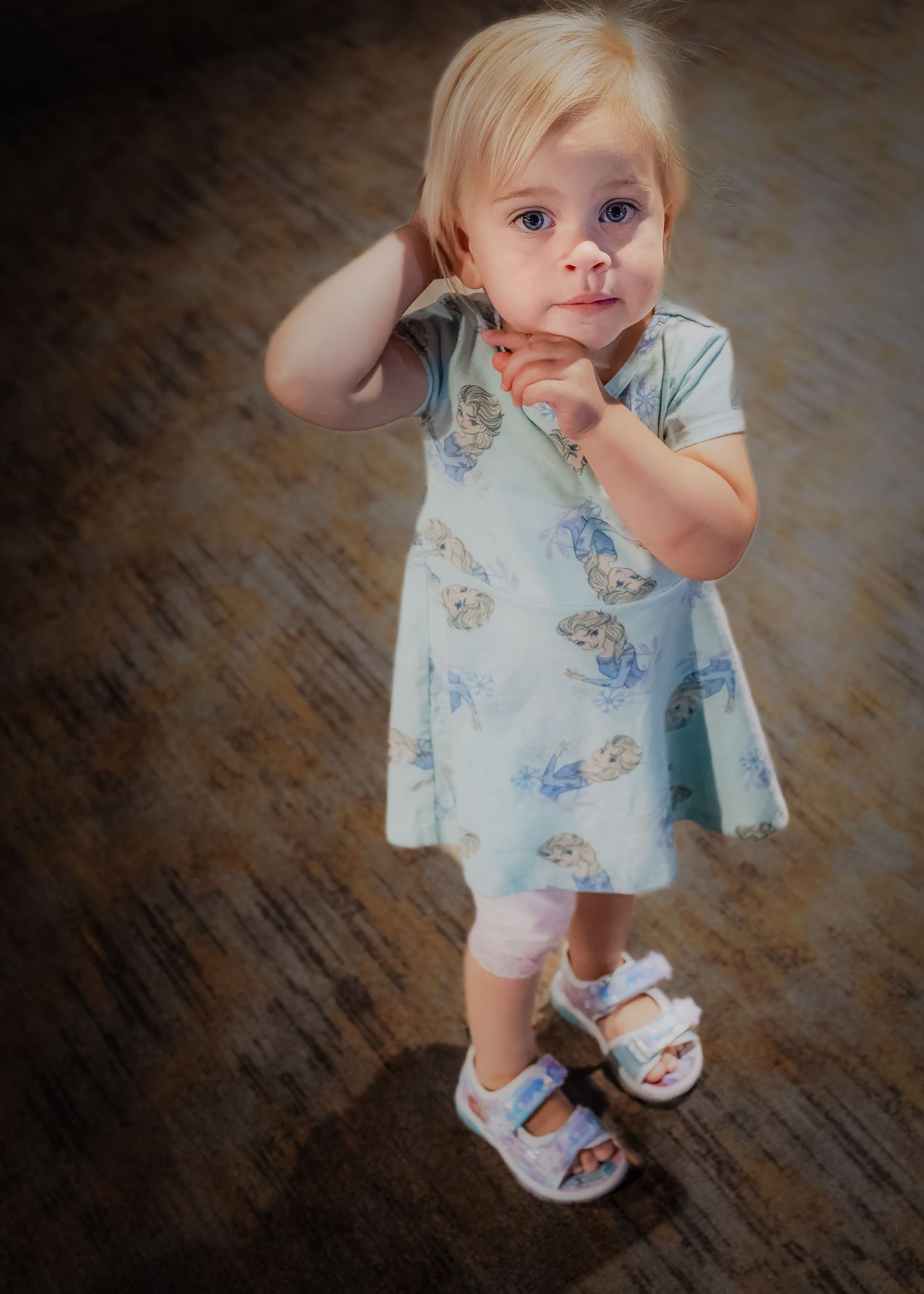 Small toddler wearing light blue floral dress and white sandals stands on dark wooden floor in sweet pose.