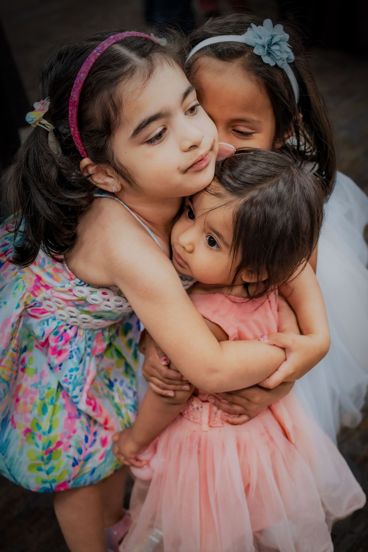 Three children sharing a tender embrace while wearing colorful dresses with hair accessories.