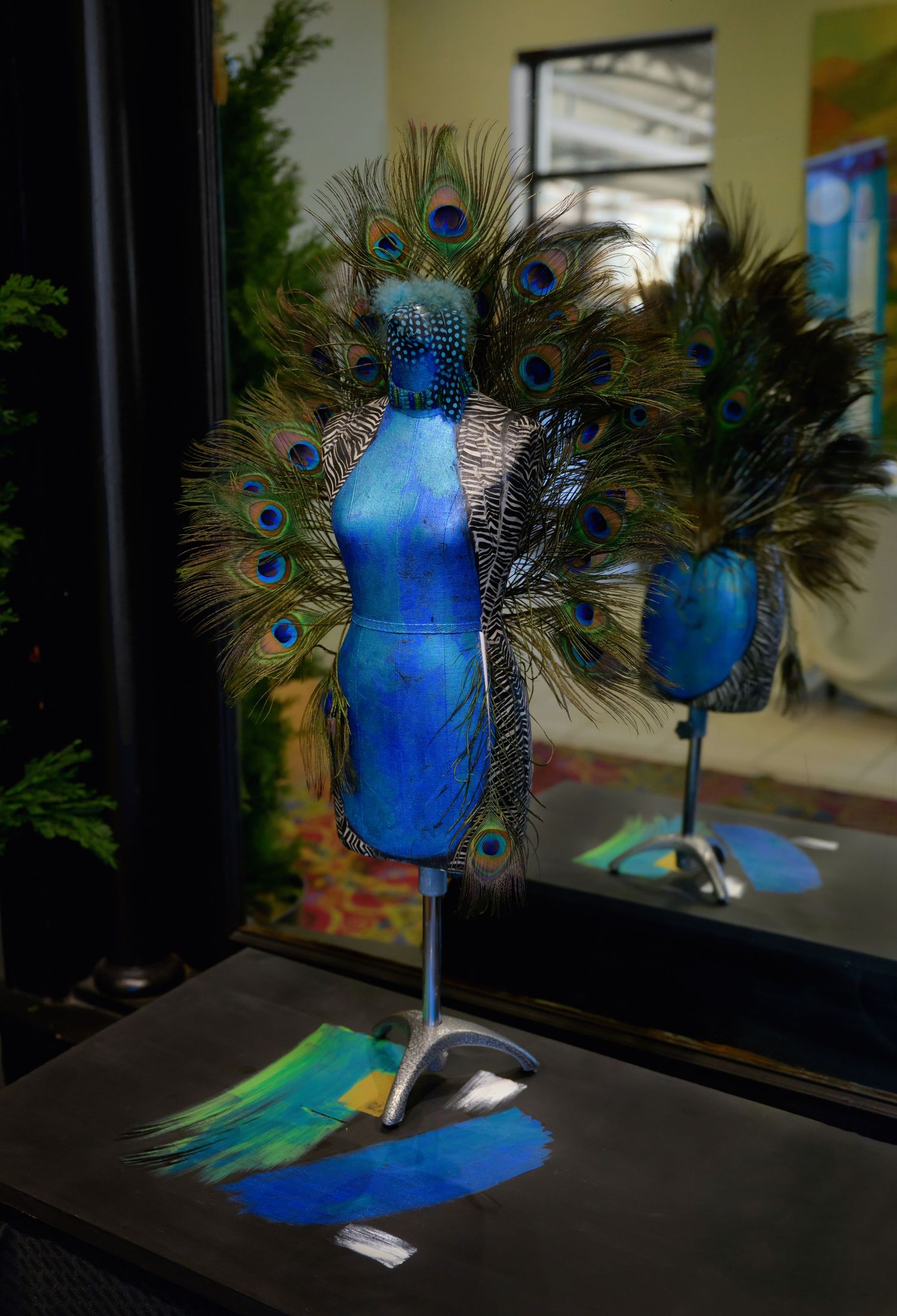 A blue dress form decorated with peacock feathers stands elegantly on a display stand with a mirror reflection.