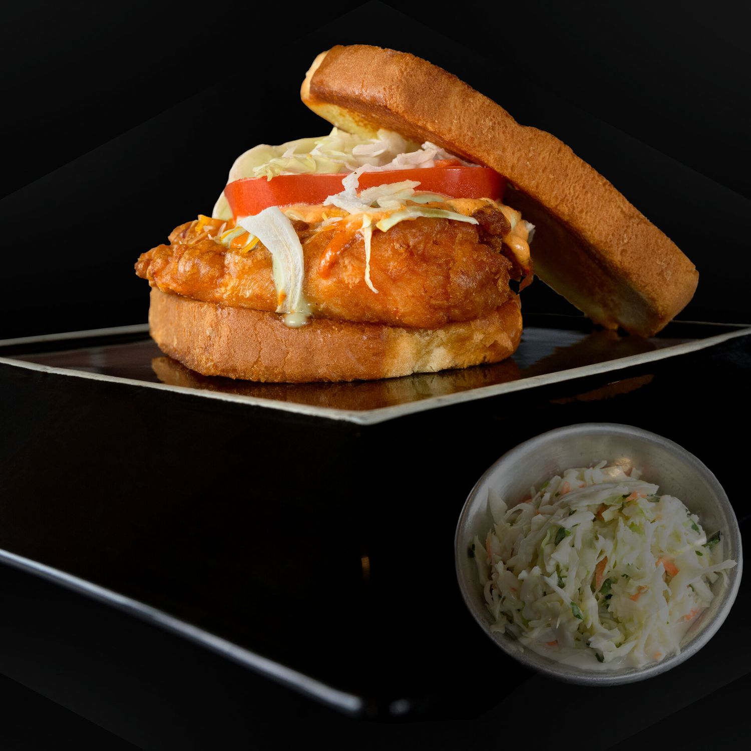 A crispy fried chicken sandwich with coleslaw and sauce on a bun, served with a side of fresh slaw.