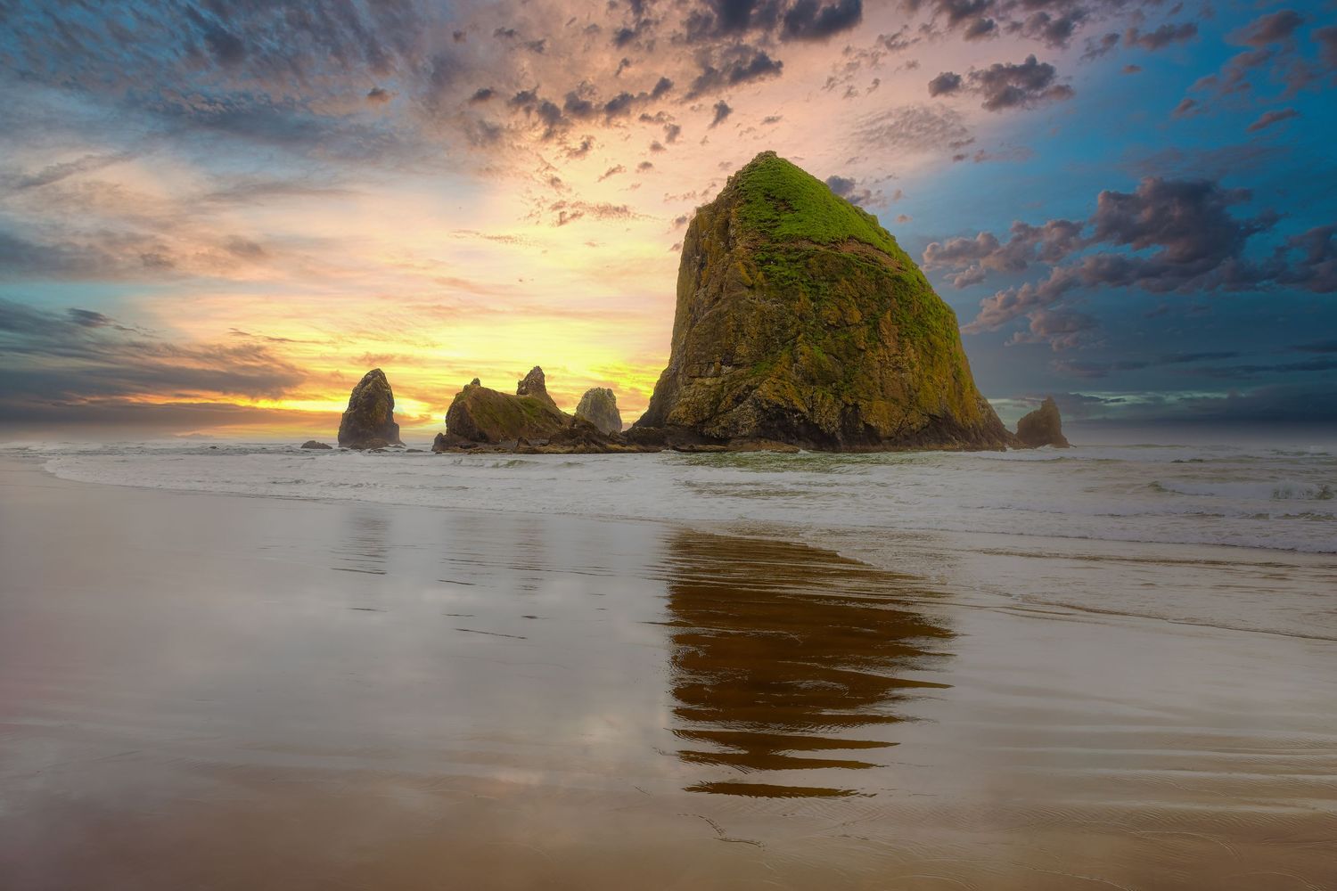 Dramatic sunset sky over Haystack Rock and needle formations at Cannon Beach, Oregon with wet sand reflections.