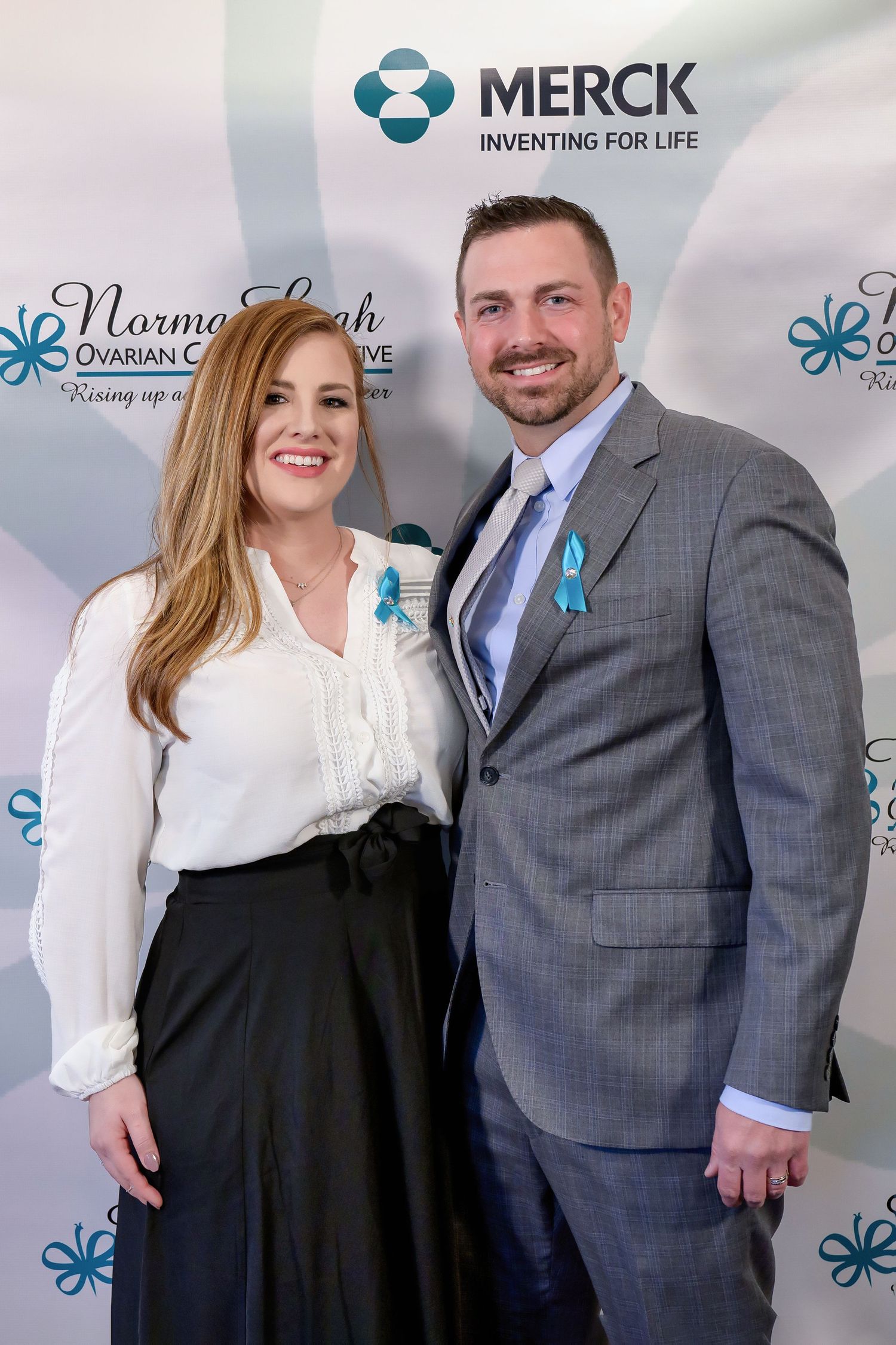 Two people in business attire pose at a Merck event with teal company logo and blue awareness ribbons.