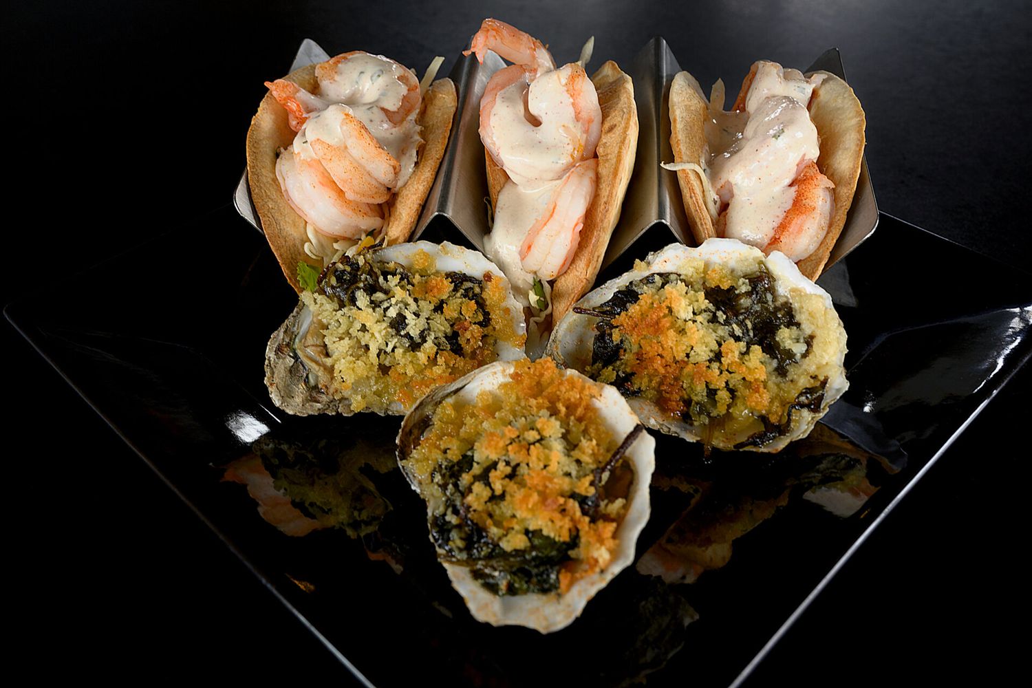 Gourmet appetizer plate featuring baked oysters with herbs and breadcrumbs alongside fresh shrimp cocktails in paper cones.
