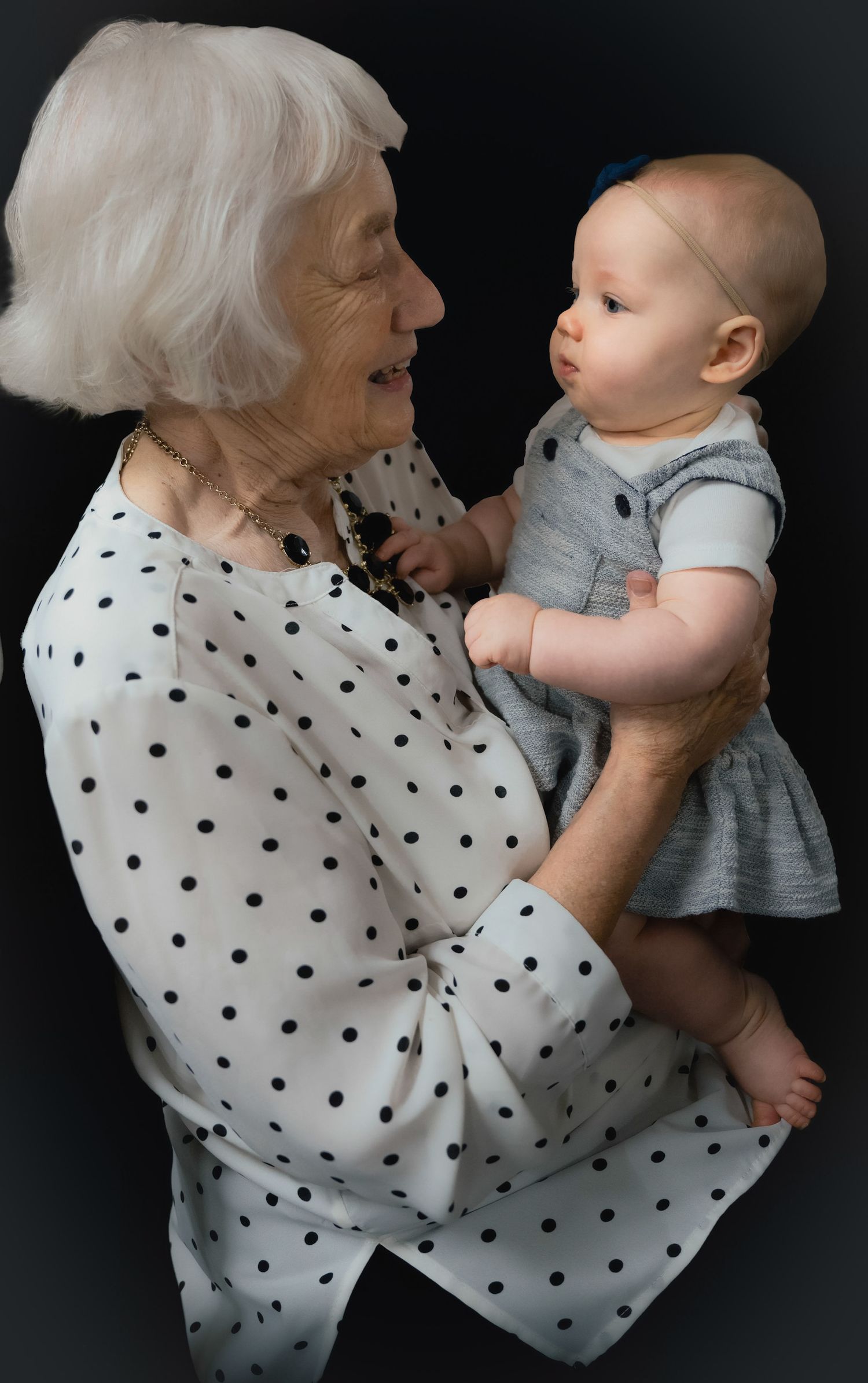 A tender moment between a white-haired person in a polka dot blouse and an infant wearing a gray dress against a dark background.