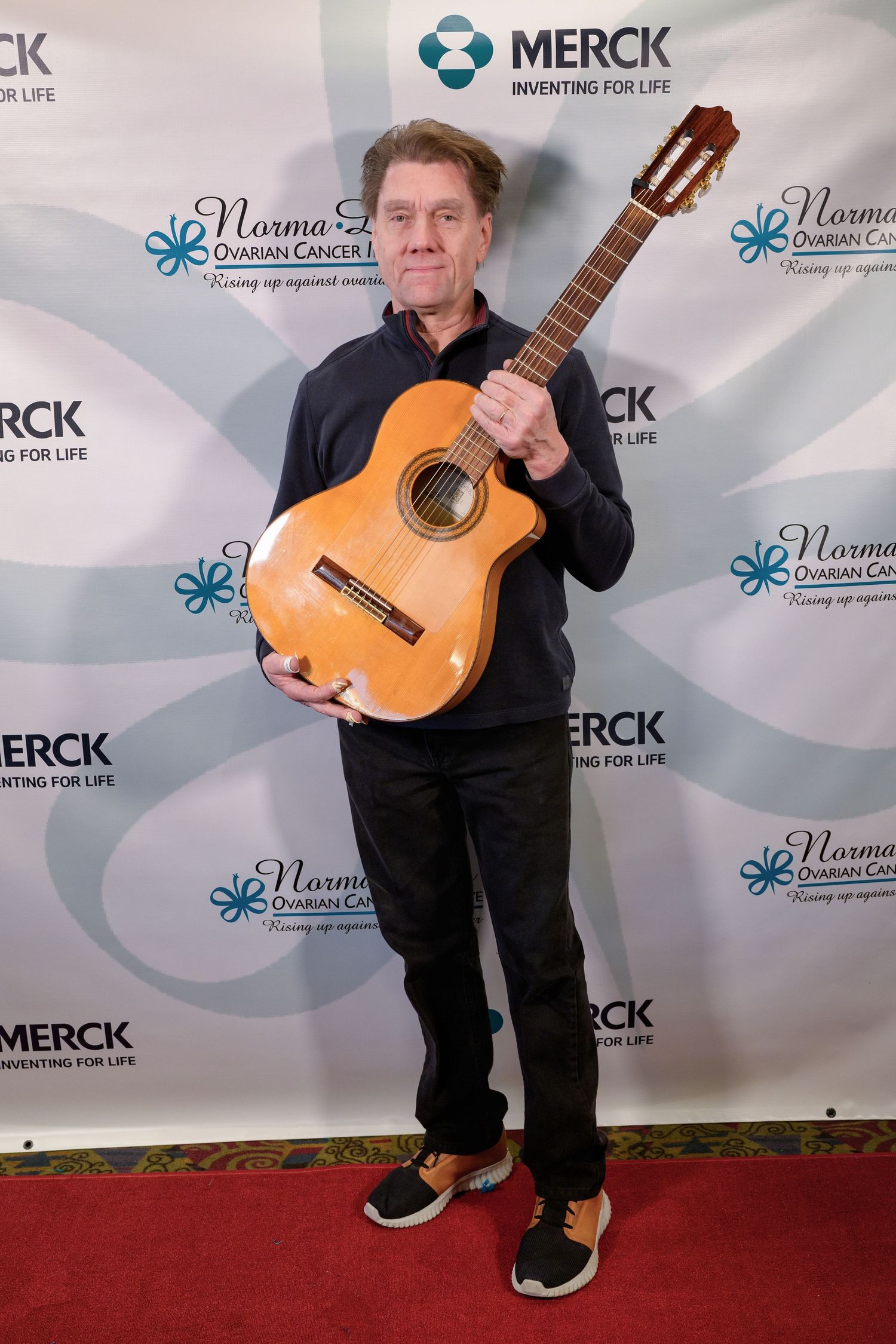 Musician stands on red carpet holding classical acoustic guitar at Merck sponsored event with branded backdrop.