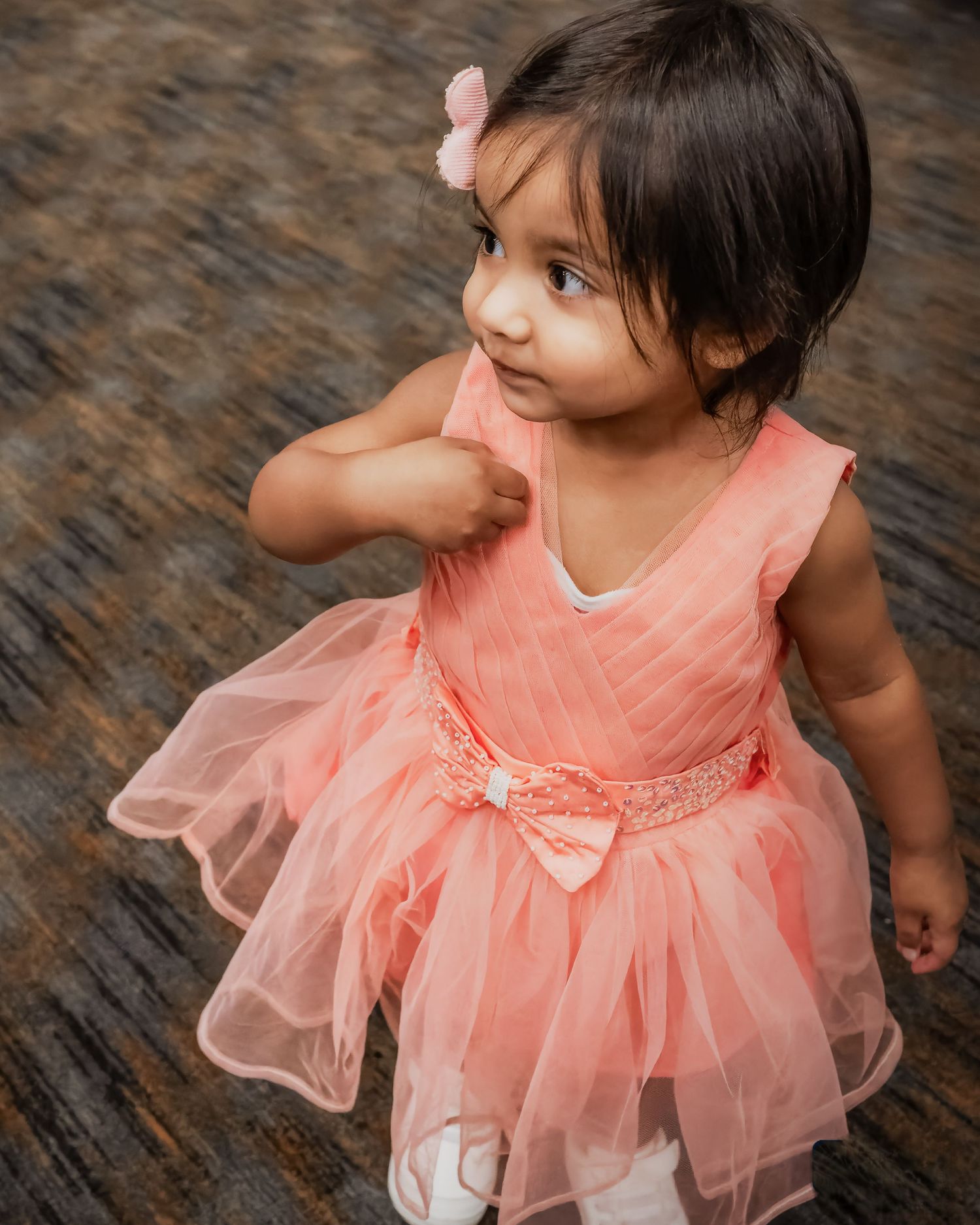 Child wearing a peach-colored tulle dress with bow detail twirls on a dark textured floor.