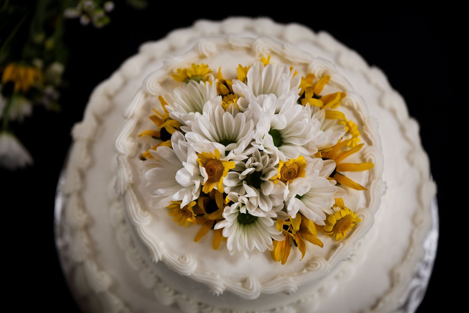 White wedding cake decorated with fresh white daisies and yellow flowers arranged in a circular pattern on top.