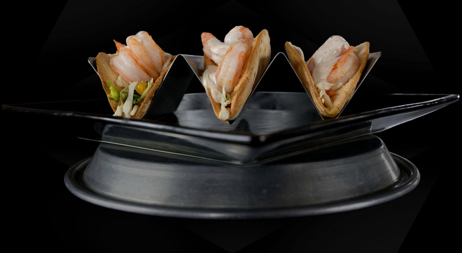Three shrimp tacos arranged on a sleek black serving platter against a dark background.