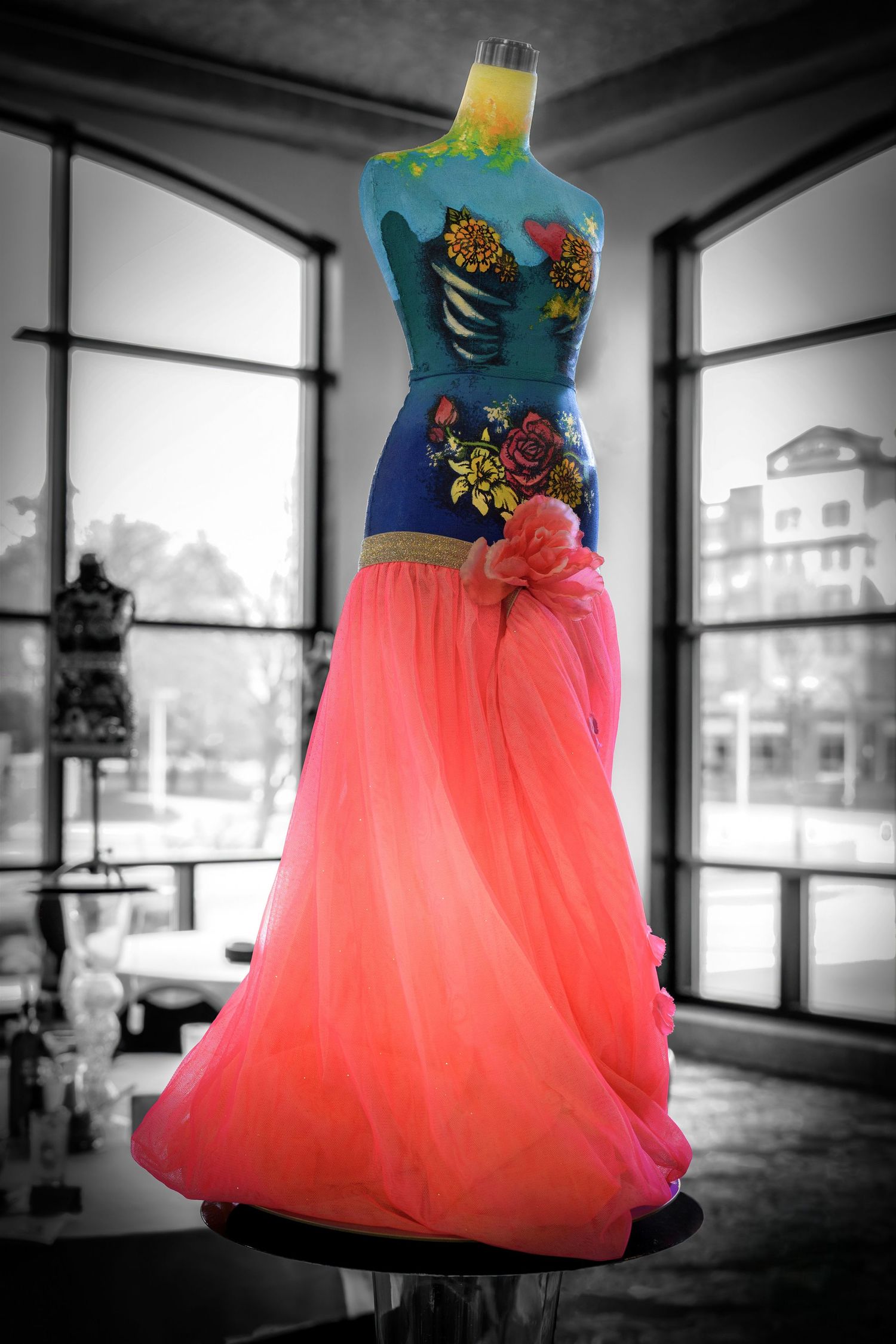 A turquoise and coral ballgown with floral embellishments stands elegantly on a dress form against a black and white background.