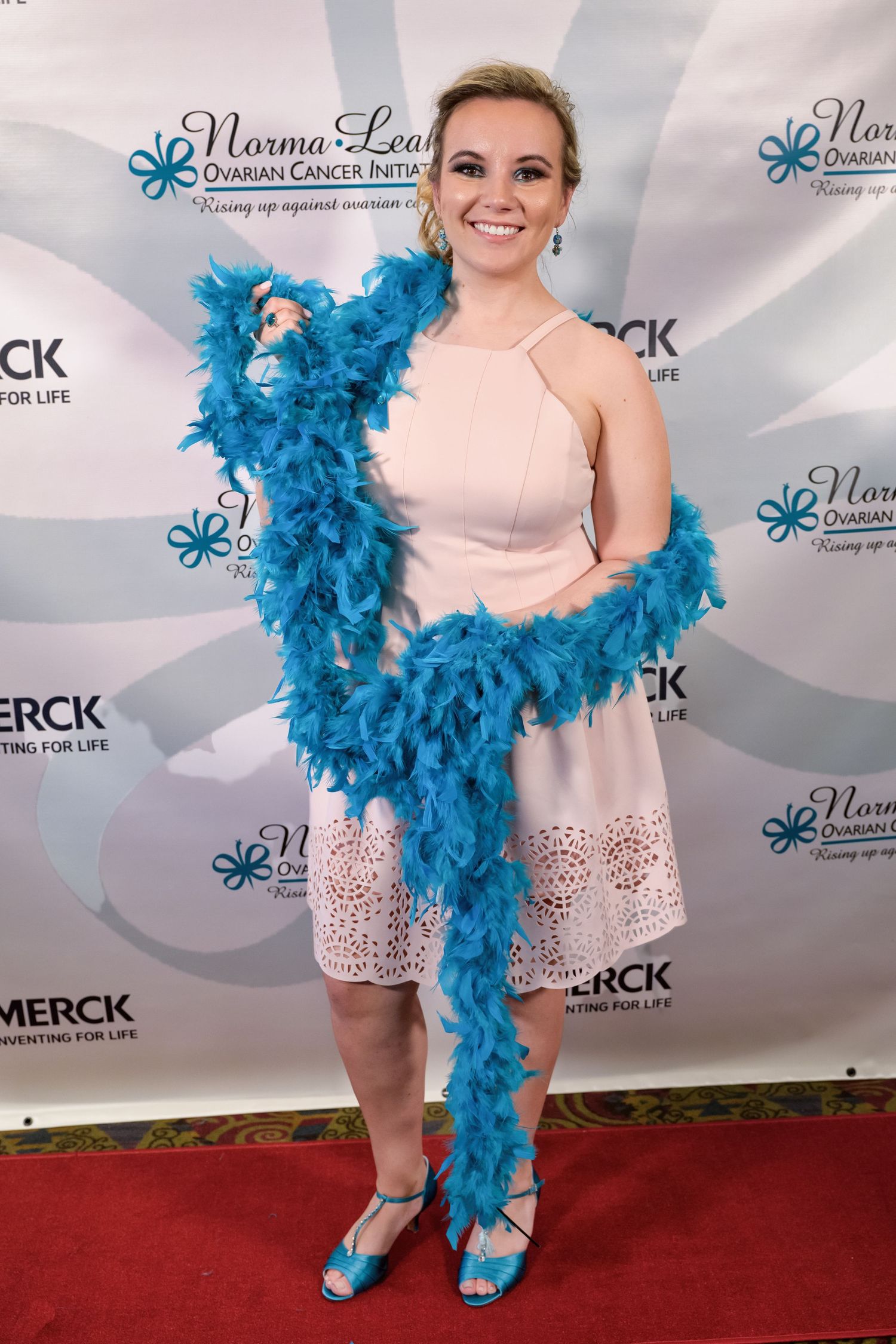 Person in pale pink dress accessorized with bright turquoise feather boa poses on red carpet at Merck event.