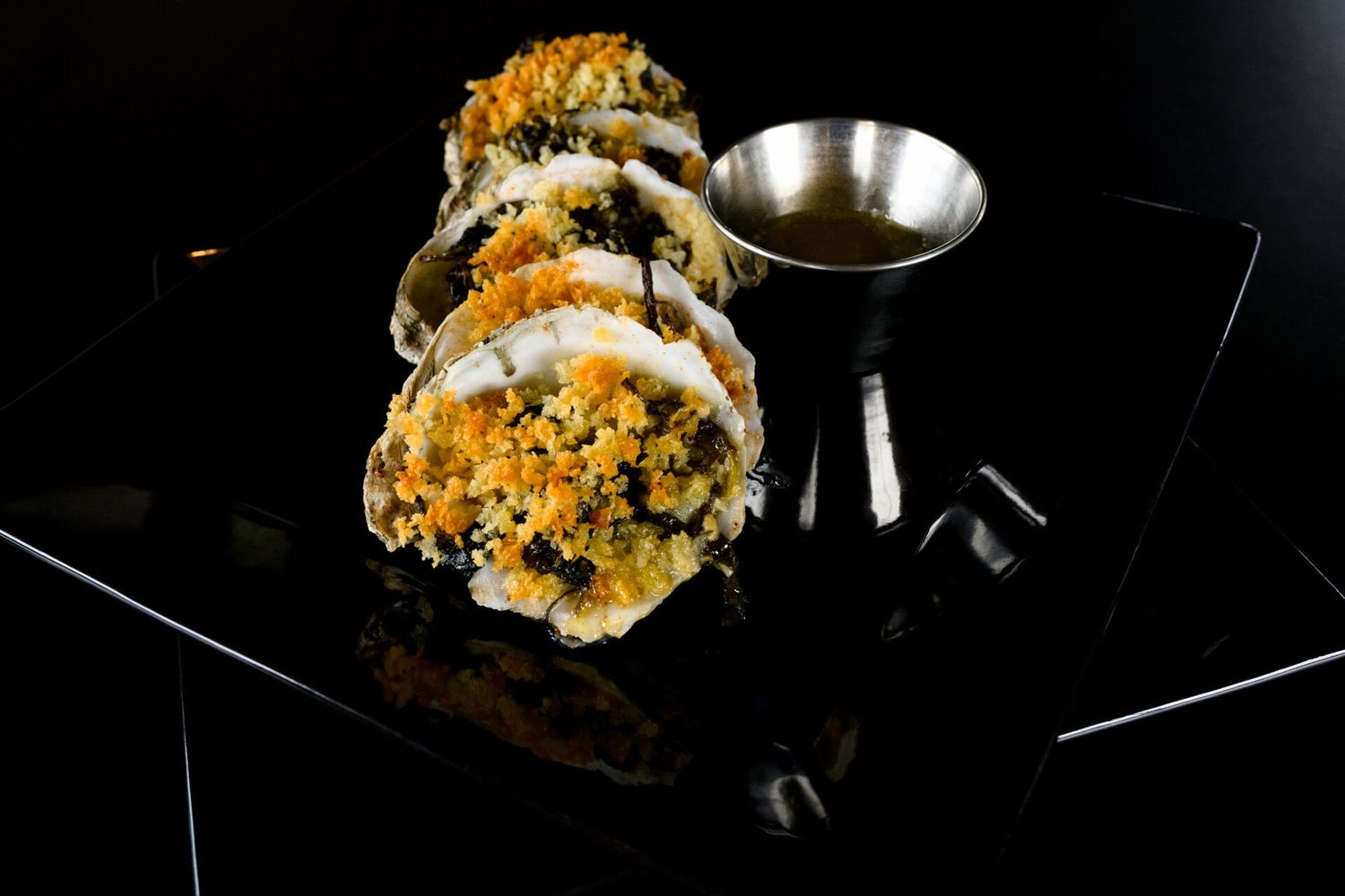 Sushi rolls topped with orange roe and herbs served on a black plate with a metal sauce cup.