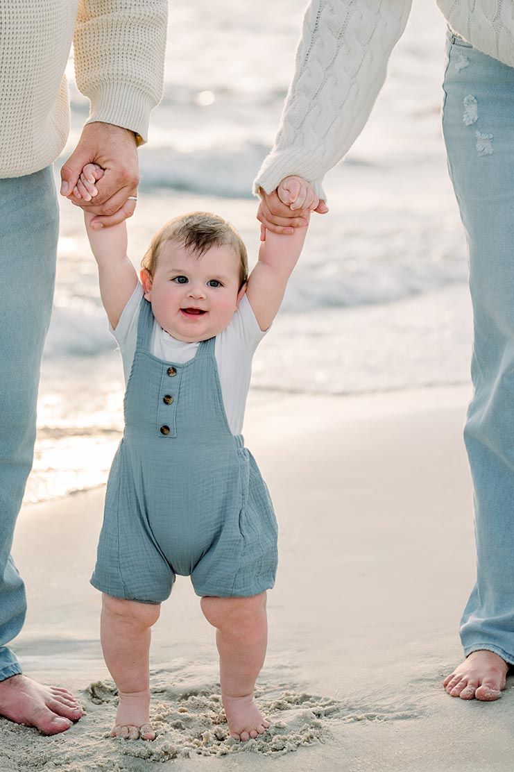 Close-up of tiny feet and legs in blue romper walking on beach sand with adult guidance.