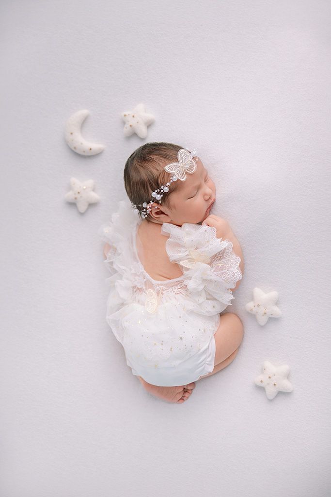 Newborn baby girl sleeping peacefully in white lace outfit with floral headband, surrounded by star and moon props.