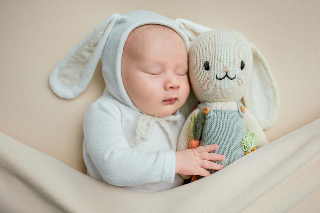 Sleeping baby in bunny hat cuddles knitted rabbit toy, wearing white outfit, peaceful newborn photography.