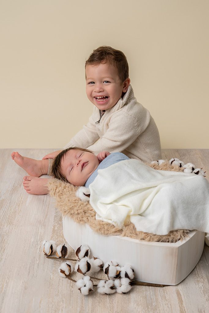 Smiling toddler boy poses with sleeping newborn baby sibling in white wooden box with cotton flowers.