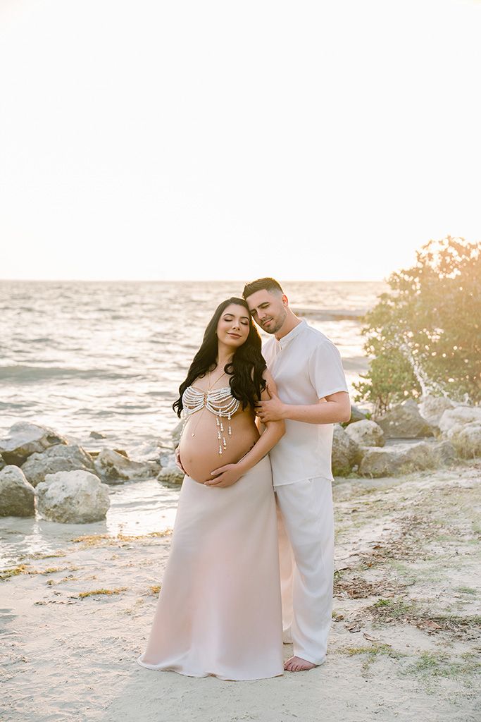 Pregnant couple poses on rocky beach at golden hour, woman in blush gown cradling bump, man in white embracing her.