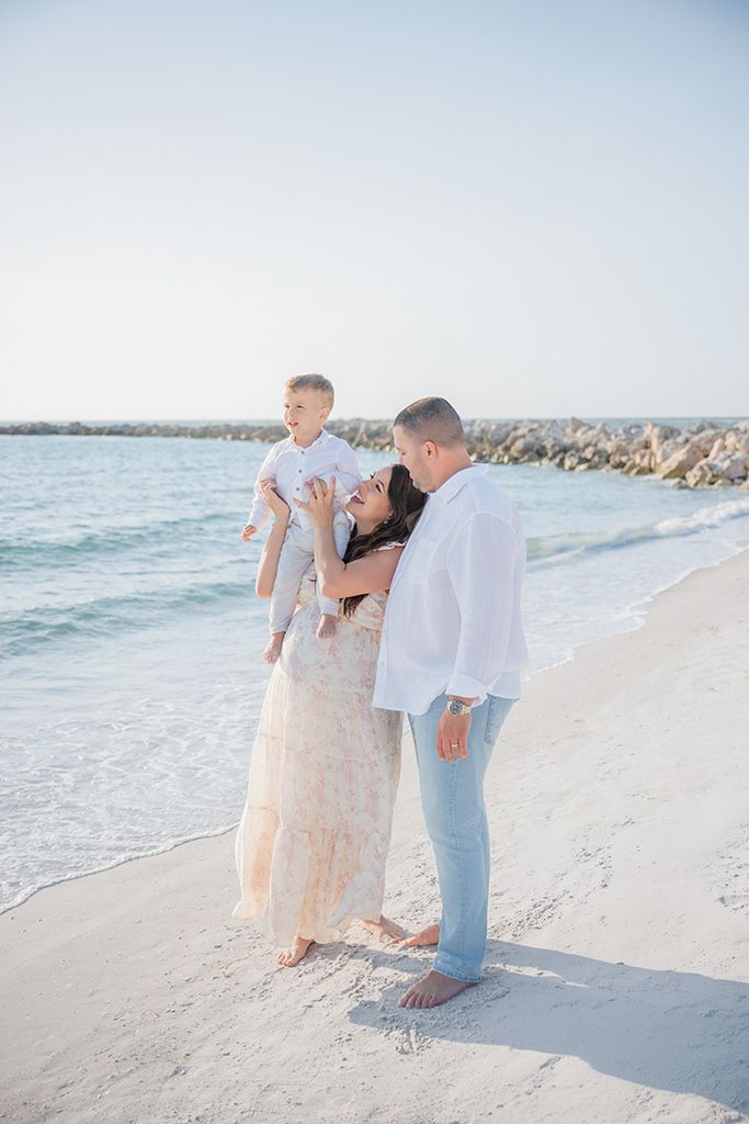 Family of three on a white sand beach, mother in lace dress with father in white shirt lifting young son.