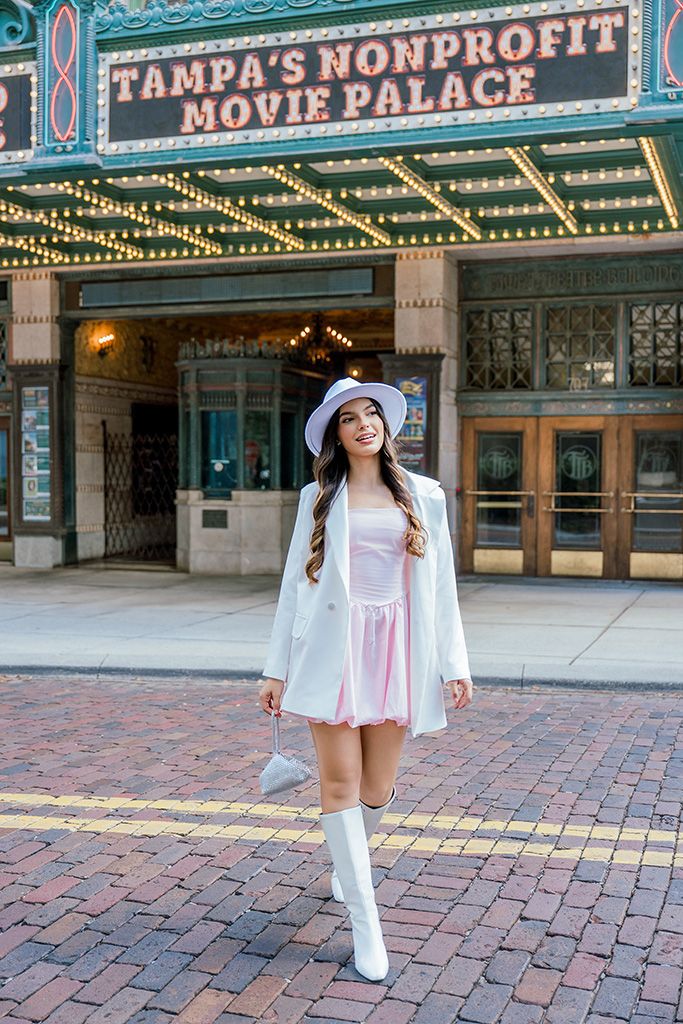 Woman in pink dress, white blazer, hat, and boots poses outside Tampa's Nonprofit Movie Palace theater.