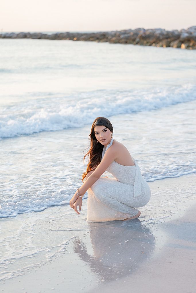 Woman in white dress crouching gracefully at the ocean's edge on a sandy beach.