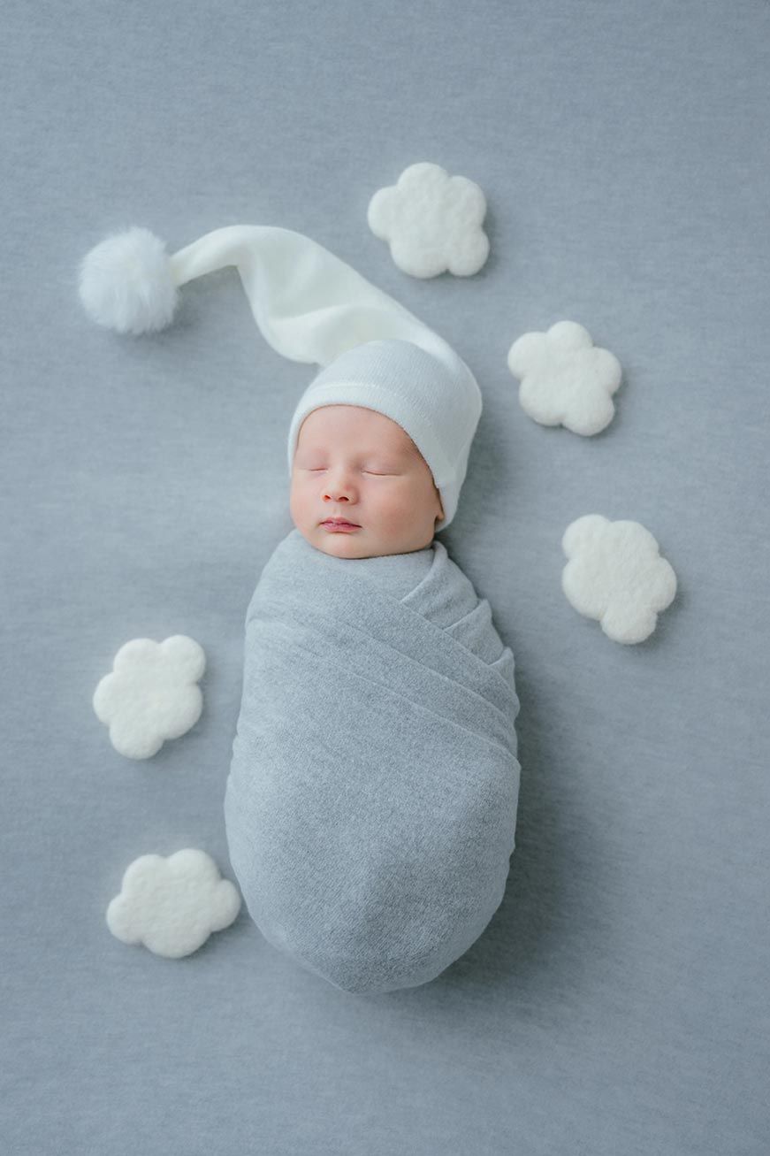 Sleeping newborn wrapped in soft gray swaddle wearing white hat surrounded by cloud-shaped cotton decorations.