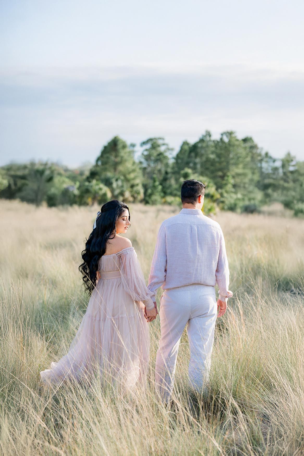 A couple holds hands while walking through tall golden grass in a romantic outdoor field setting at sunset.