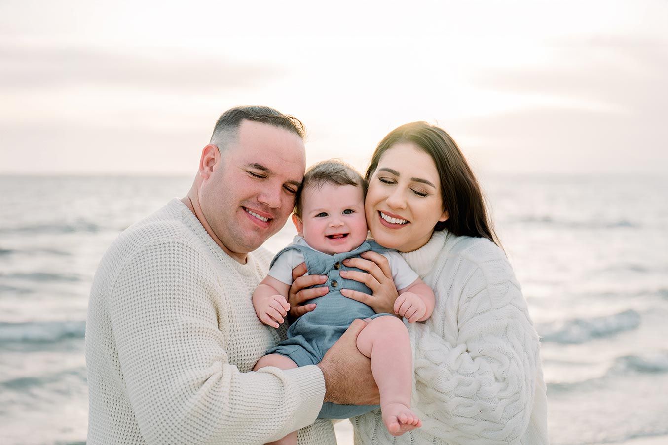 Happy family with baby in blue romper posing together on a beach at sunset.