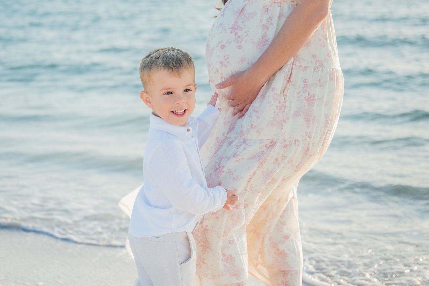 Young boy in white hugs pregnant mother in floral dress on sunny beach.