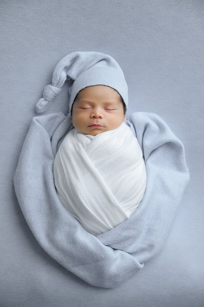Peaceful newborn baby swaddled in white wrap, wearing a gray knit hat, sleeping on a soft blue blanket.