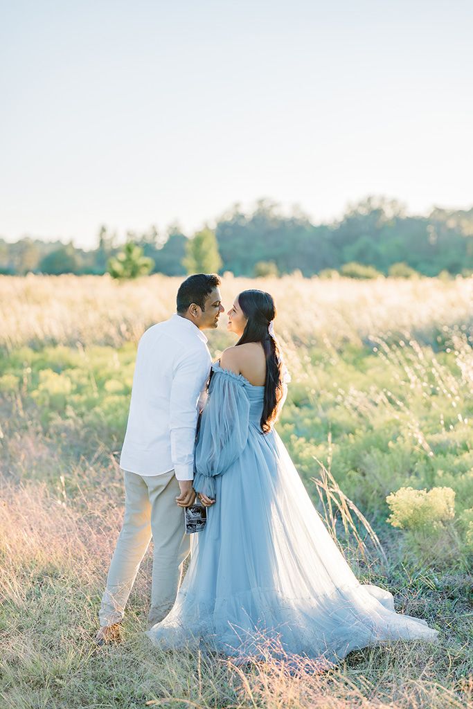 A couple shares a kiss in a golden field, the woman wearing an elegant blue tulle gown, holding a sonogram photo.