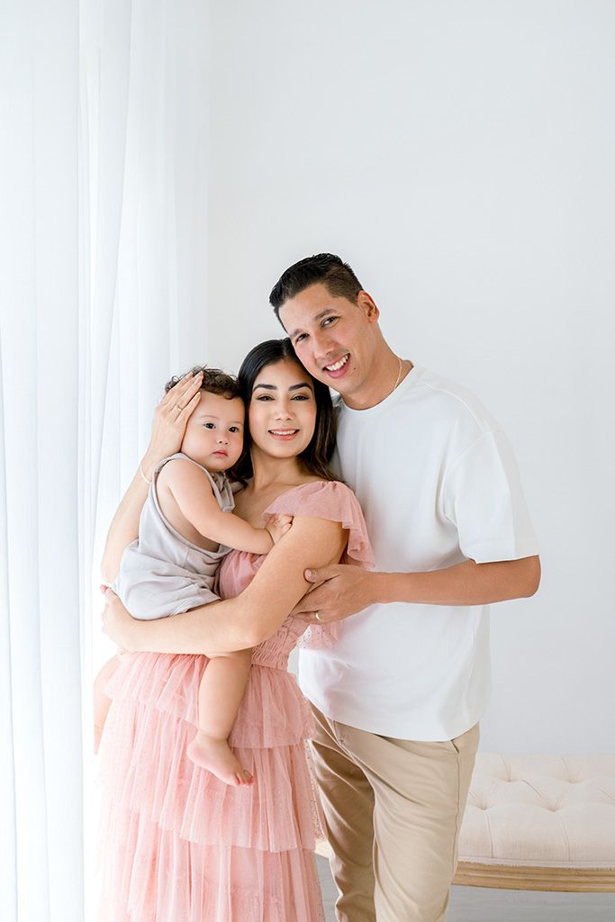 Happy Asian family portrait with mother in pink dress holding toddler, father in white tee, posing in bright white room.