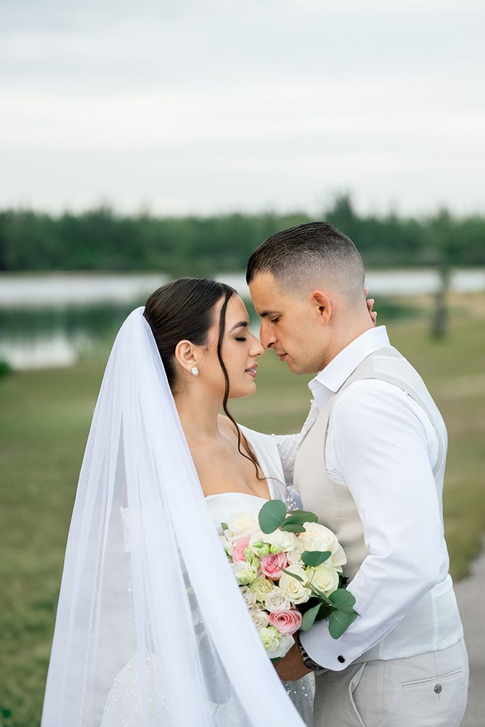 Bride and groom share an intimate moment outdoors, with white veil and pink rose bouquet near a lake.