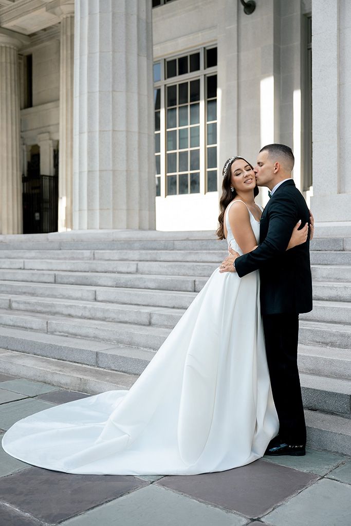 Bride in white gown with train and groom in black tuxedo embrace on marble steps of elegant building.