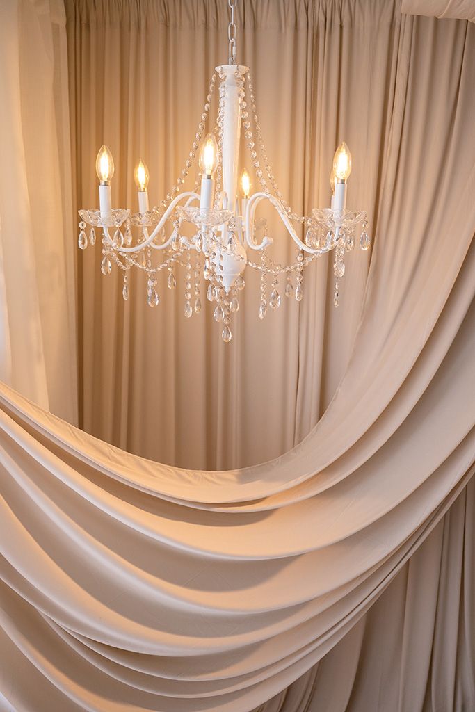 Elegant white crystal chandelier with candle lights hanging against draped beige satin curtains.