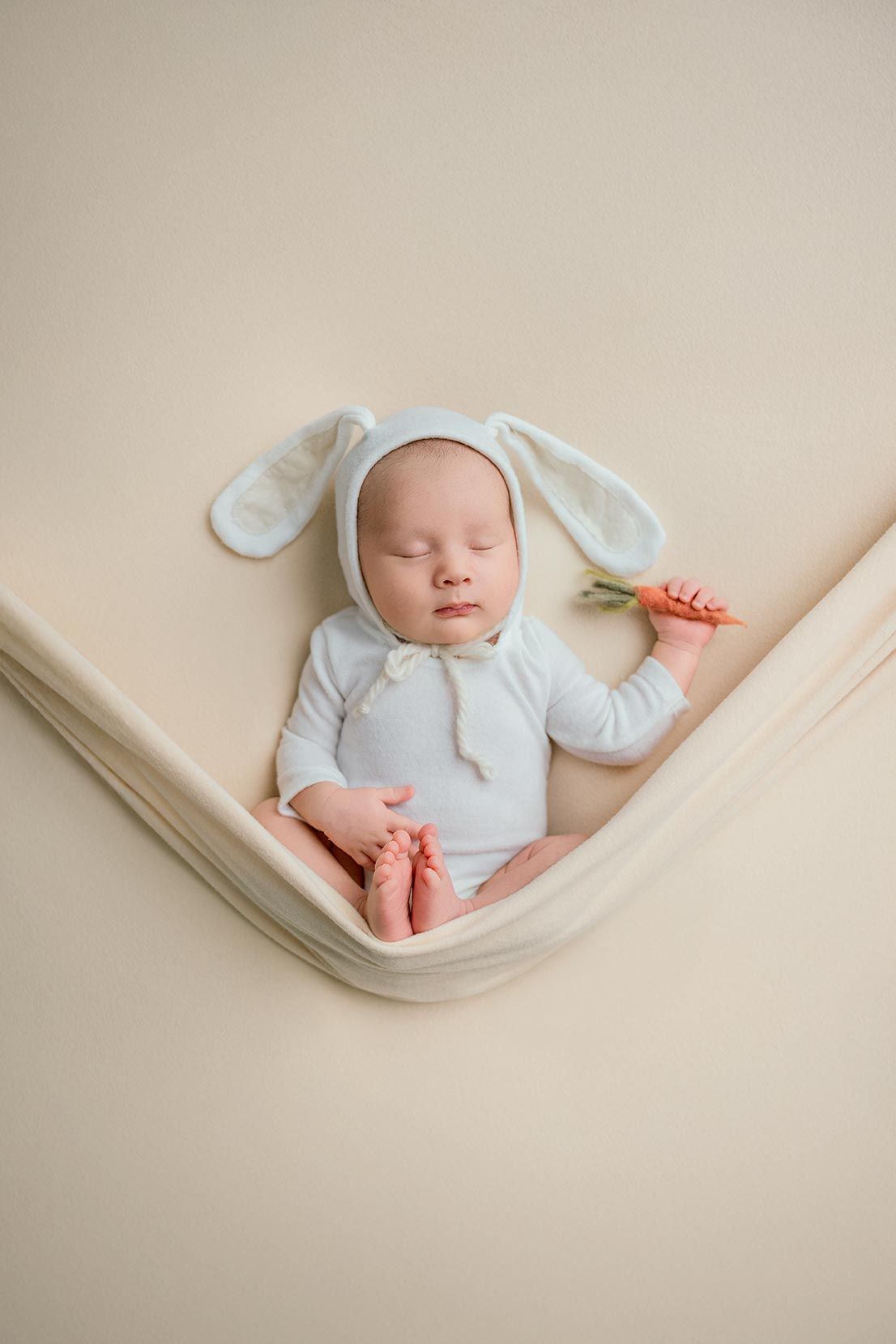 Sleeping newborn wearing white bunny ears outfit lies peacefully on cream-colored fabric background.