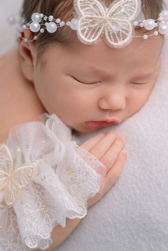 Sleeping newborn baby girl wearing white lace outfit and floral pearl headband, close-up portrait.