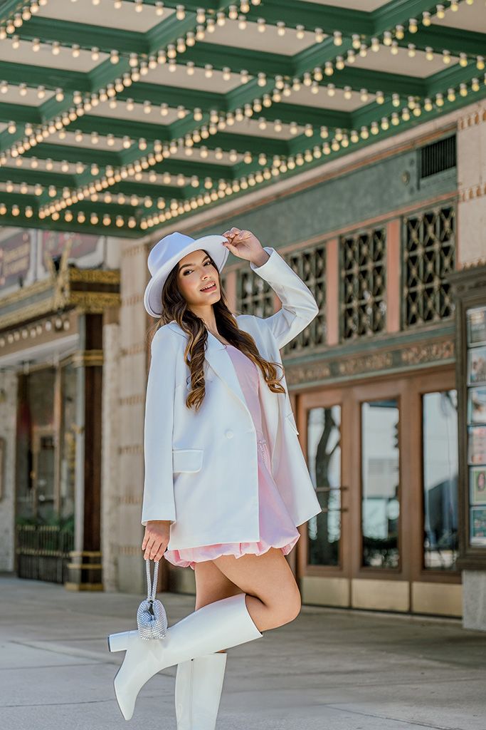 Stylish woman wearing white blazer, pink mini dress, knee-high boots and fedora hat at theater.
