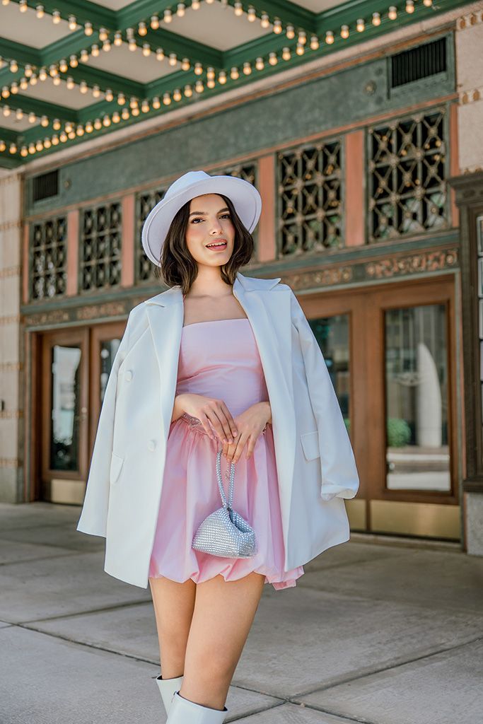 Young woman in pink mini dress, white blazer and bucket hat holds small bag outside decorative theater.