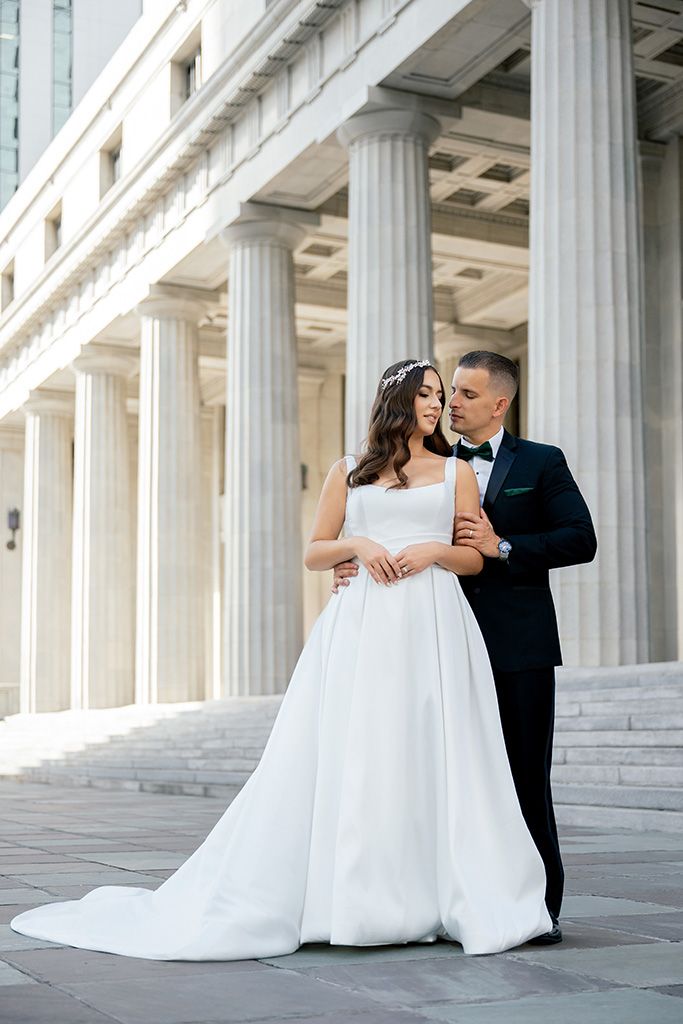 Bride and groom embrace before grand white columns on wedding day, bride in elegant white gown with train.