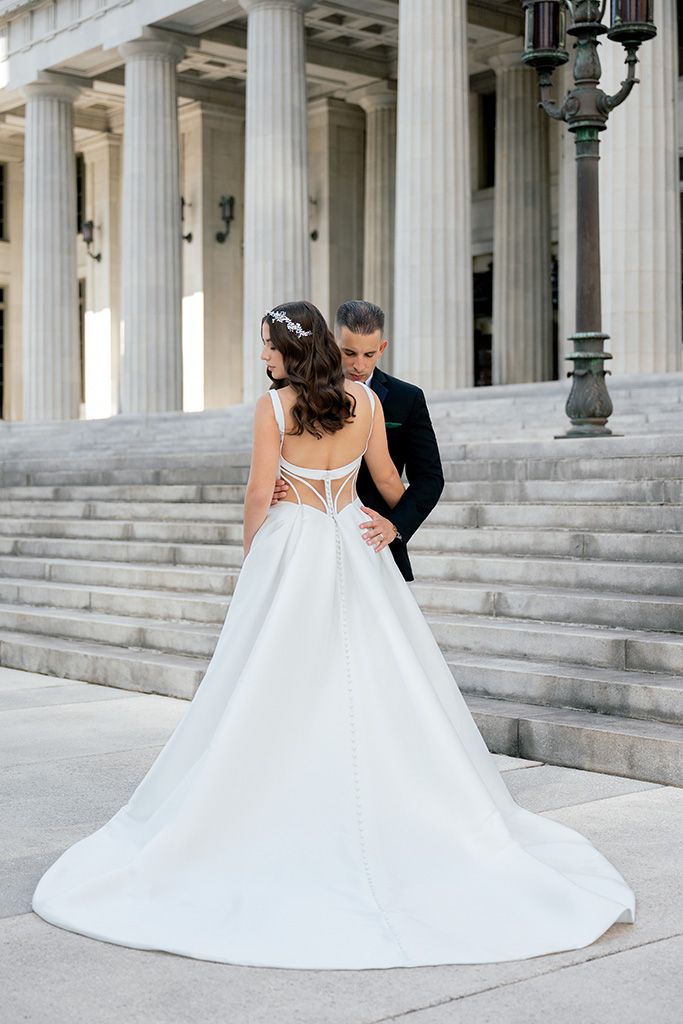 Bride in elegant white gown with strappy back stands with groom in black tux on stone steps of classic building.