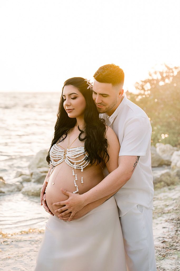 Pregnant couple embracing at golden hour beach, woman in pearl bodychain with flowing skirt, man in white shirt holding her belly.