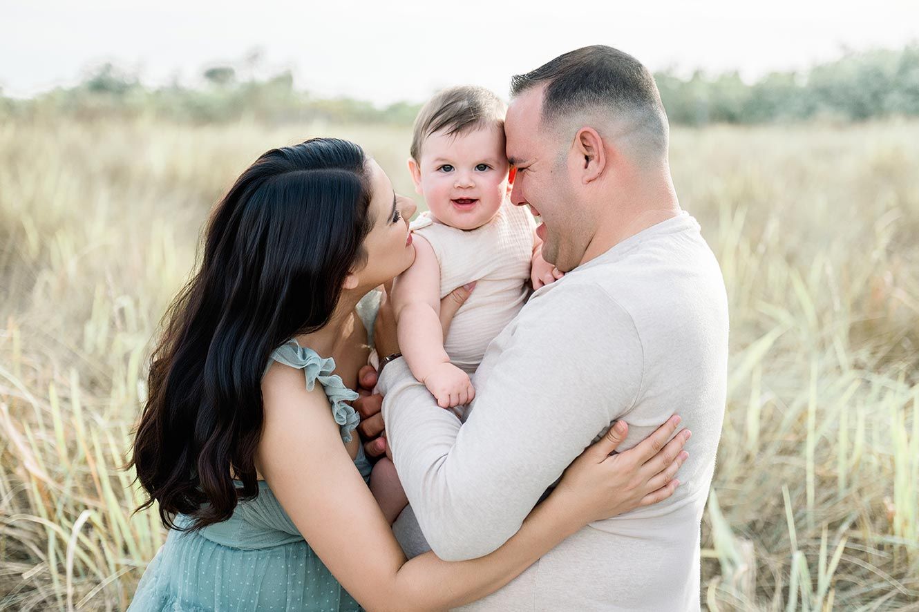 Parents kissing baby girl in a sunlit field, family portrait photography session outdoors.