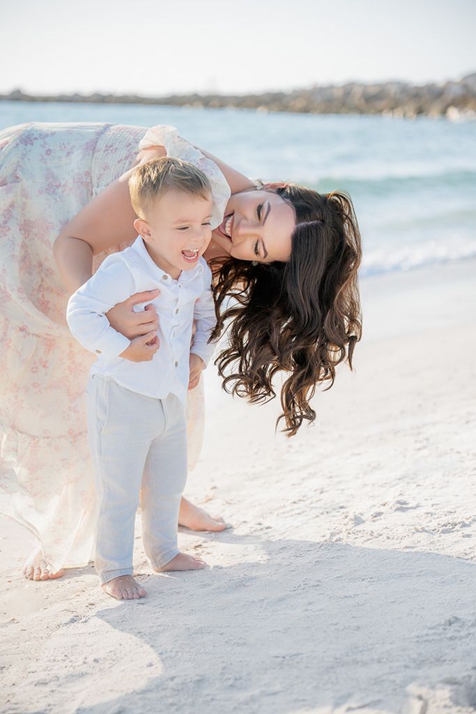 Mother kissing toddler son on beach, both dressed in white and pastels, with ocean waves and white sand.