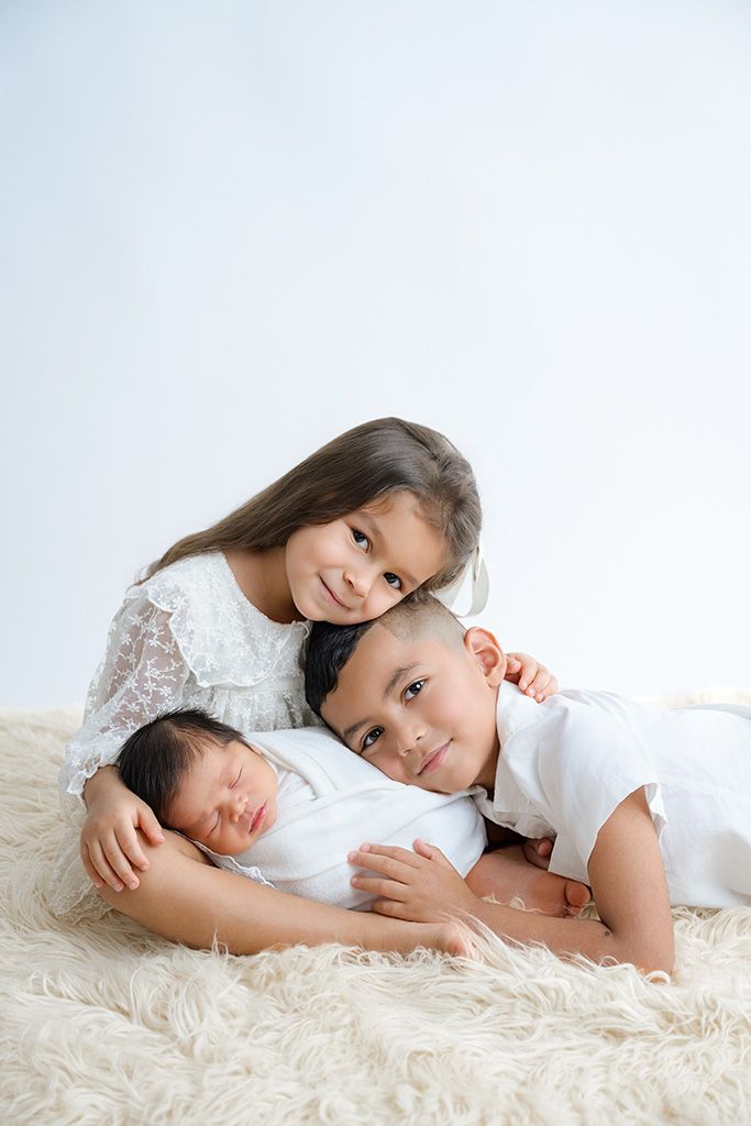 Two older siblings in white outfits lovingly pose with their sleeping newborn baby on a fluffy cream rug.