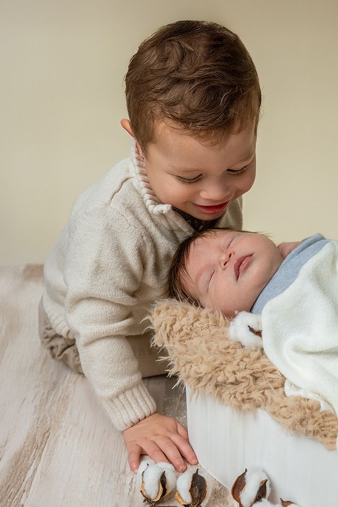 A young toddler boy in a beige sweater lovingly leans over a sleeping newborn baby wrapped in a soft blanket.