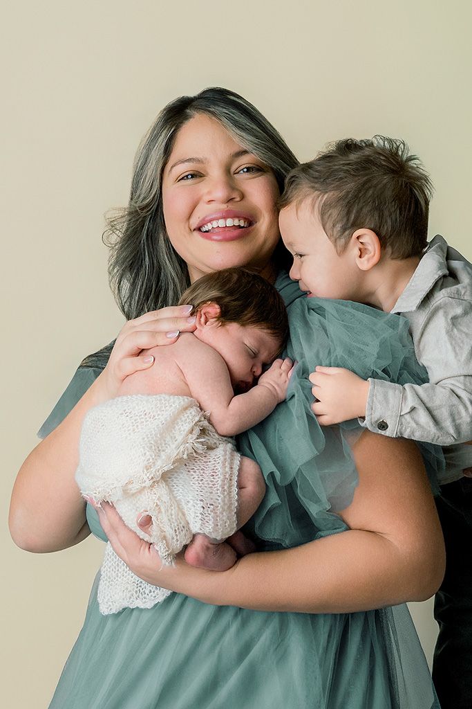 Smiling mother in teal dress holds newborn baby while young toddler son leans in for a kiss on neutral background.