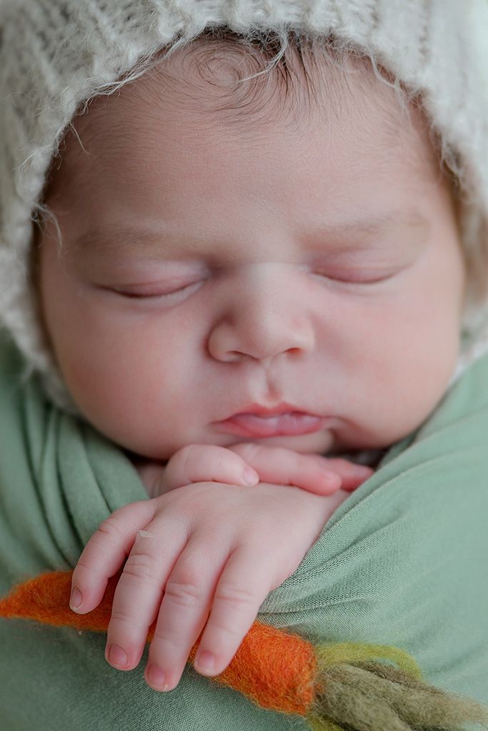 Close-up of sleeping newborn baby wearing knit hat, wrapped in green swaddle holding a carrot prop.
