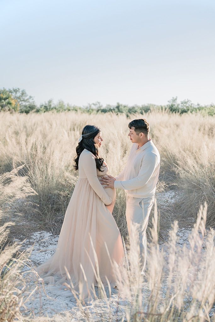 Pregnant couple embracing in golden field, woman in blush gown, man in white, sharing tender moment outdoors.