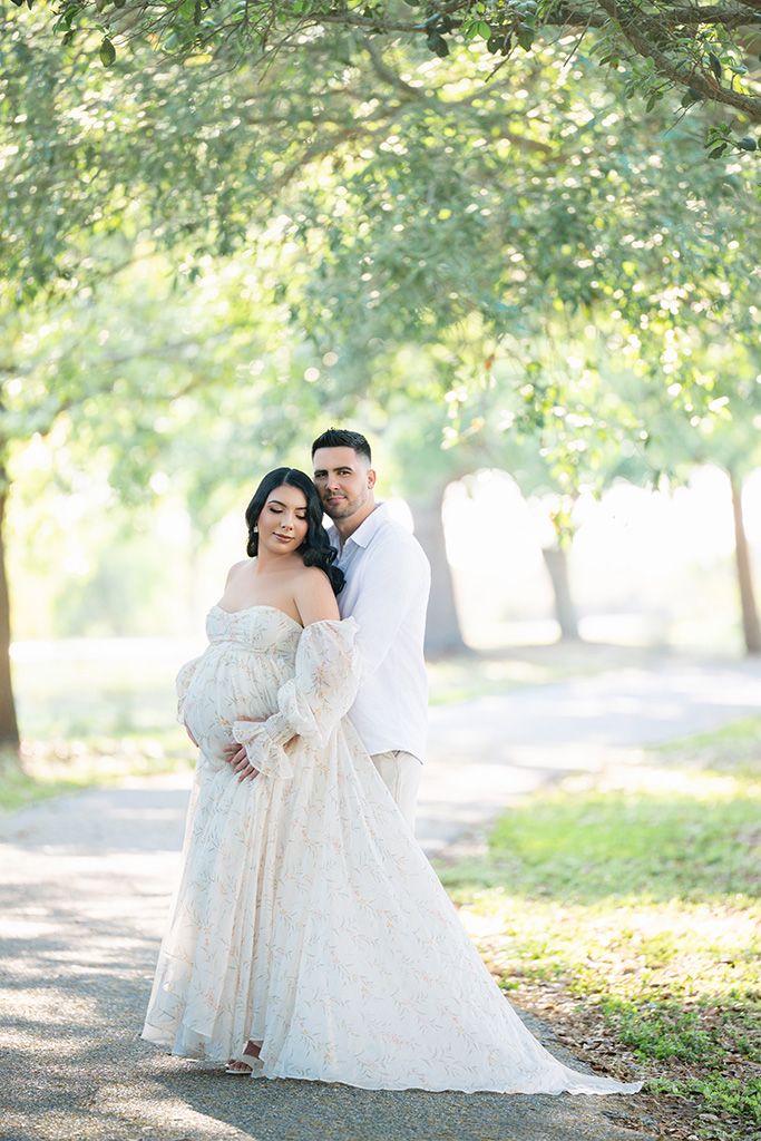 Couple poses on a tree-lined path, pregnant woman in lace gown embraced by man in white shirt.
