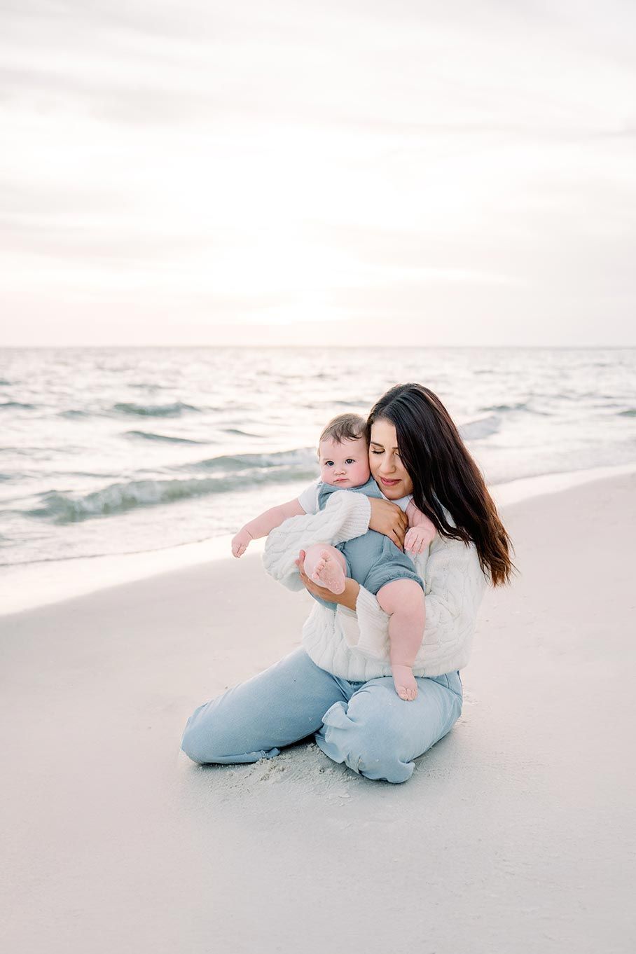 A mother sits on a white sand beach holding her baby, with gentle ocean waves in the background at sunset.