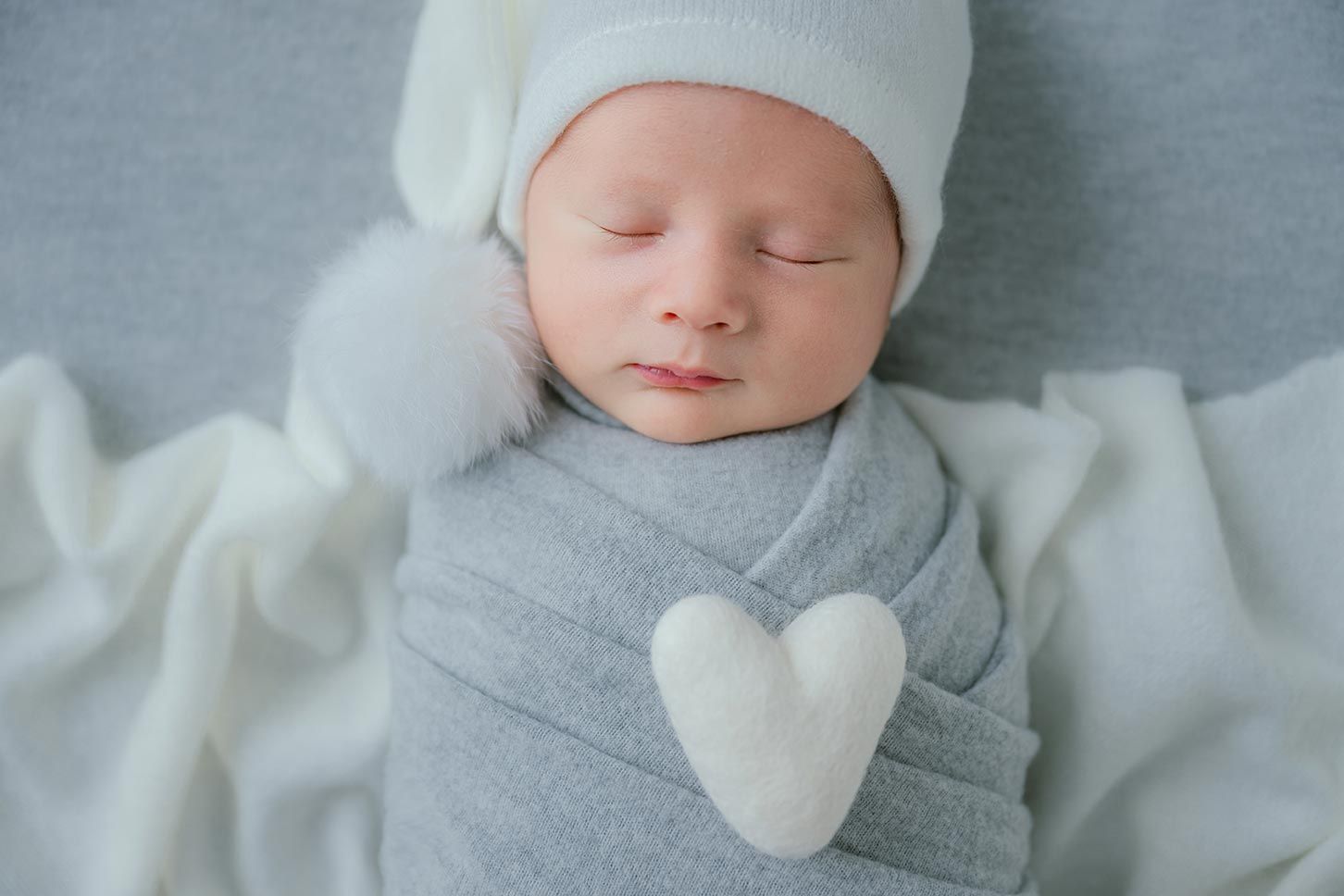 Sweet newborn baby in gray outfit with white bow tie sleeping peacefully on soft white blanket.